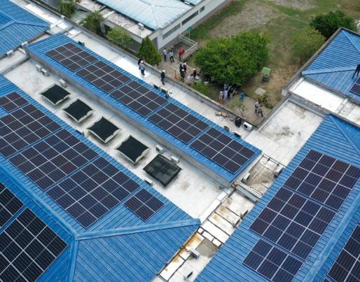 The image shows an aerial view of a building with several solar panels installed on its blue roofs. In the top right corner, a group of people appears to be observing or inspecting the installation.