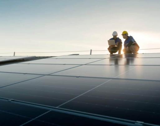 Two workers wearing helmets inspect solar panels at sunset. The scene shows a solar energy facility undergoing technical maintenance.