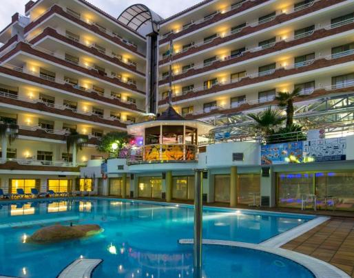 Multi-storey hotel lit up at dusk, featuring a large pool and lounging area. Palm trees, reclining chairs, and an elevated structure complete the holiday atmosphere