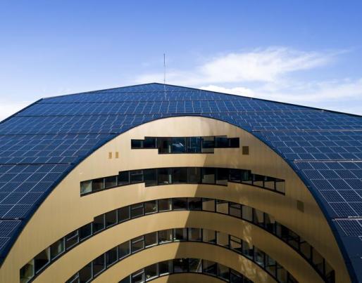 The image shows a modern building with a curved façade and aligned windows. The roof is covered with solar panels, indicating the use of solar energy.