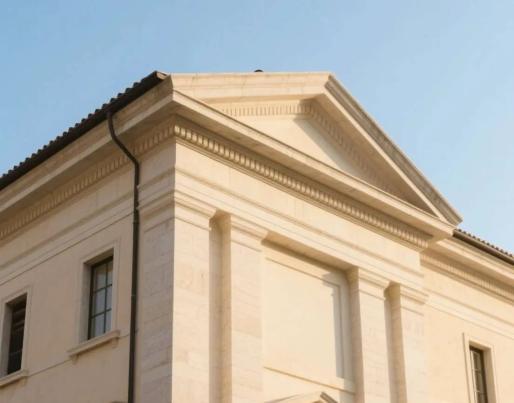 Upper part of a classical-style building with a triangular pediment, columns, and rectangular windows. The light-coloured façade stands out against the clear blue sky.