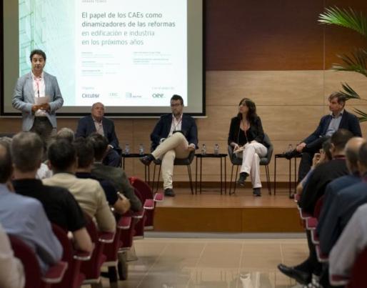 A conference with five speakers on stage and an attentive audience. The screen highlights the role of CAEs in building and industry reforms.