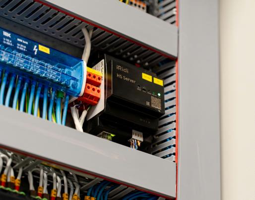 The image shows an electrical control panel with various components and wiring. A blue device labelled "iRidi HS Server" stands out, along with other electronic modules connected by numerous coloured wires.