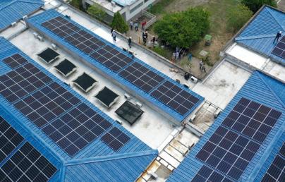 The image shows an aerial view of a building with several solar panels installed on its blue roofs. In the top right corner, a group of people appears to be observing or inspecting the installation.