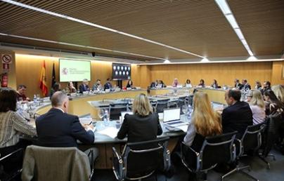 The image shows a work meeting in a modern conference room, with several people seated around an oval table. The screen displays the logo of the Government of Spain.
