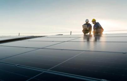 Two workers wearing helmets inspect solar panels at sunset. The scene shows a solar energy facility undergoing technical maintenance.