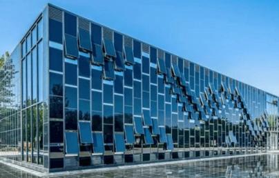 The image shows a modern building with a reflective glass façade and a geometric design. It is surrounded by trees, and a pond in front mirrors its structure.