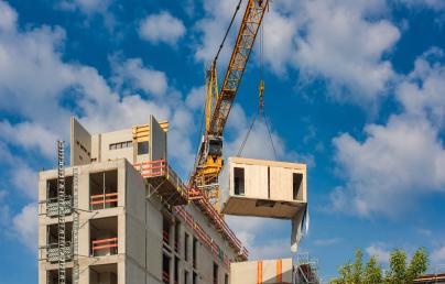 A prefabricated module is being lifted by a crane onto a multi-storey building under construction. The site is active under a clear sky with scattered clouds, showcasing offsite construction in progress.