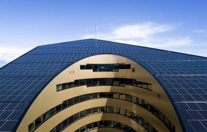 The image shows a modern building with a curved façade and aligned windows. The roof is covered with solar panels, indicating the use of solar energy.