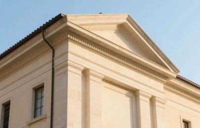 Upper part of a classical-style building with a triangular pediment, columns, and rectangular windows. The light-coloured façade stands out against the clear blue sky.