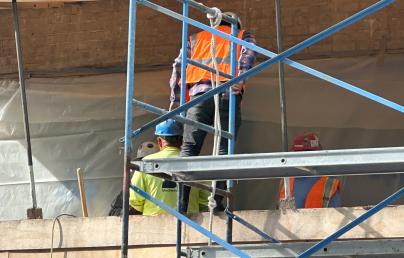 A group of construction workers in reflective vests and helmets are working on scaffolding in front of a brick wall. The metal structure of the scaffolding is prominent in the foreground.