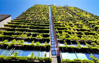 The image shows a tall building with a unique architectural design, featuring a façade covered in plants that creates a vertical garden effect. This highlights sustainable practices and urban greening.