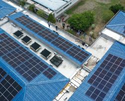 The image shows an aerial view of a building with several solar panels installed on its blue roofs. In the top right corner, a group of people appears to be observing or inspecting the installation.