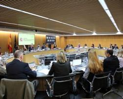 The image shows a work meeting in a modern conference room, with several people seated around an oval table. The screen displays the logo of the Government of Spain.