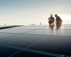 Two workers wearing helmets inspect solar panels at sunset. The scene shows a solar energy facility undergoing technical maintenance.