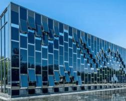 The image shows a modern building with a reflective glass façade and a geometric design. It is surrounded by trees, and a pond in front mirrors its structure.