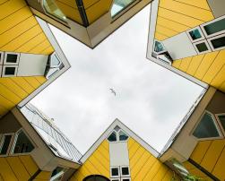 A group of yellow buildings with white windows seen from below, forming a star shape. A seagull flies in the centre of the cloudy sky.