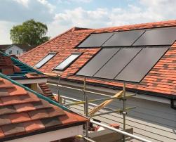 The image shows house roofs with red tiles and solar panels on one of them. There are scaffolds around, suggesting construction or maintenance work is underway.