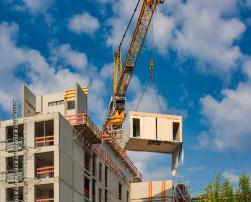 A prefabricated module is being lifted by a crane onto a multi-storey building under construction. The site is active under a clear sky with scattered clouds, showcasing offsite construction in progress.