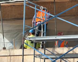 A group of construction workers in reflective vests and helmets are working on scaffolding in front of a brick wall. The metal structure of the scaffolding is prominent in the foreground.